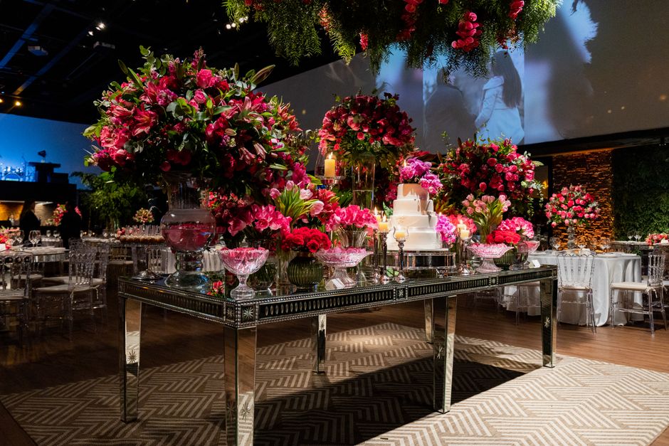 A luxurious wedding reception table decorated with an opulent arrangement of vibrant pink and deep red flowers, including roses and lilies, in elegant glass vases. The floral display features lush gre