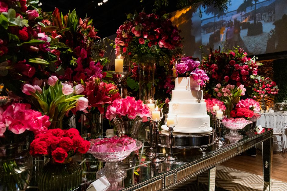 A lavish floral display featuring an array of fresh pink and red flowers such as roses, carnations, and lilies arranged in various glass vases and containers on a reflective metallic table. Tall flowe