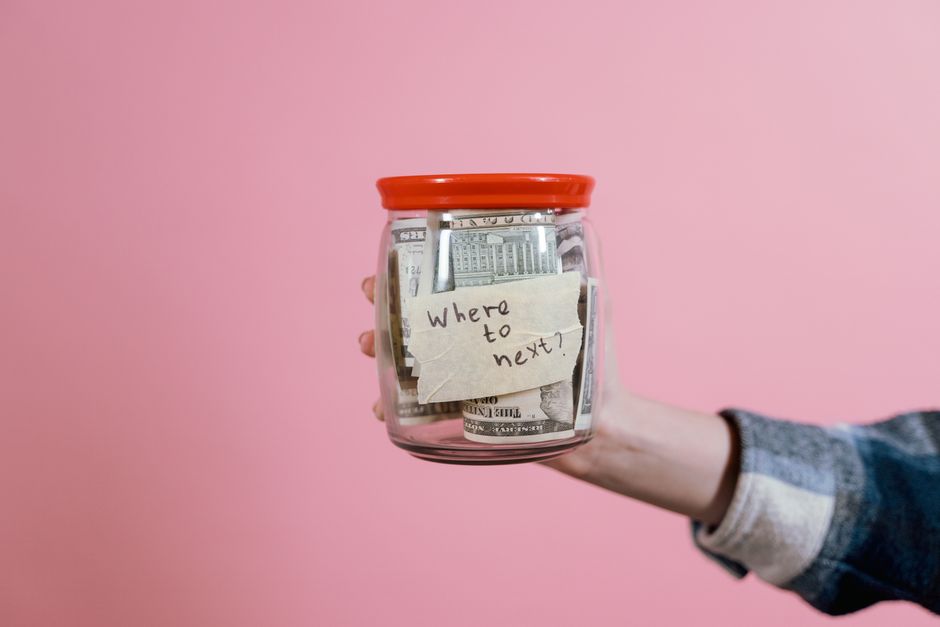 A person's hand holding a transparent glass jar filled with crumpled US dollar bills, with a small paper note attached to the jar's front stating 'where to next?'. The person is wearing a long-sleeved
